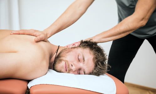 Mężczyzna leży na brzuchu na na stole fizjoterapeutycznym. Fizjoterapeuta manipuluje jego stawem barkowym, aby rozluźnić napięcie.
